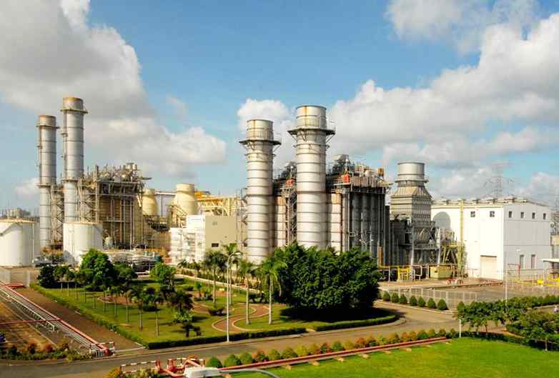 A power plant featuring tall smokestacks, industrial structures, and green landscaping under a cloudy blue sky.