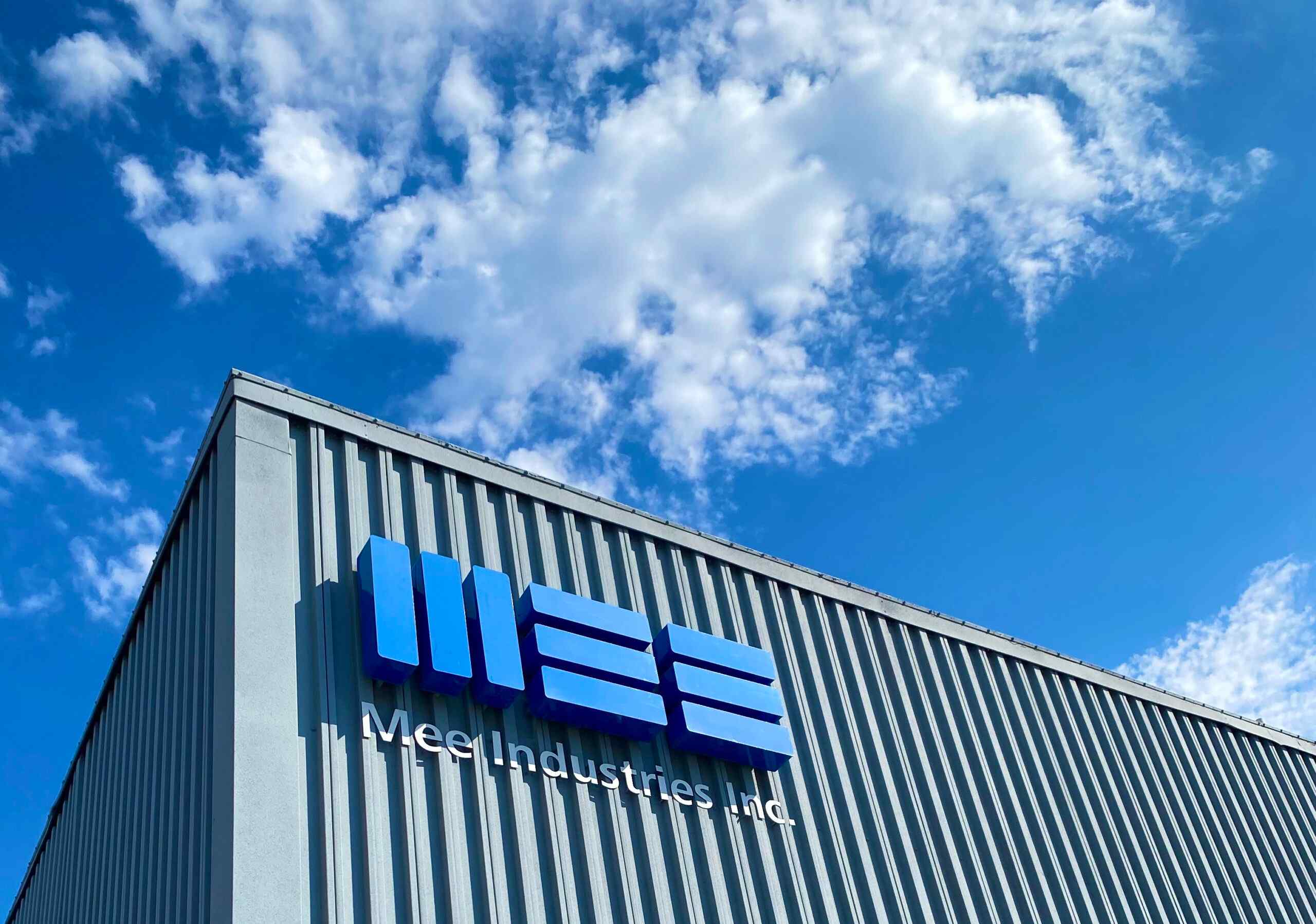 Exterior of Mee Industries building featuring a prominent blue logo against a clear blue sky with scattered clouds.