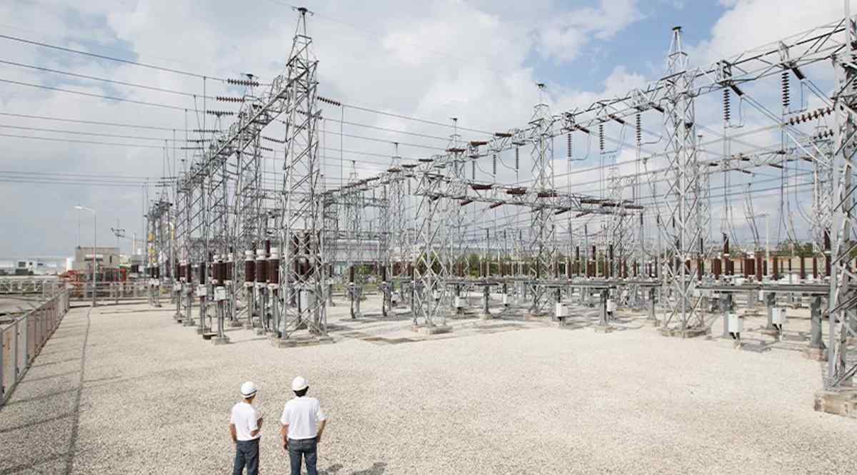 Two workers in hard hats observe a large electricity substation with metal structures and power lines against a cloudy sky.