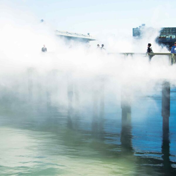 People walking through cooling mist on a waterfront bridge.