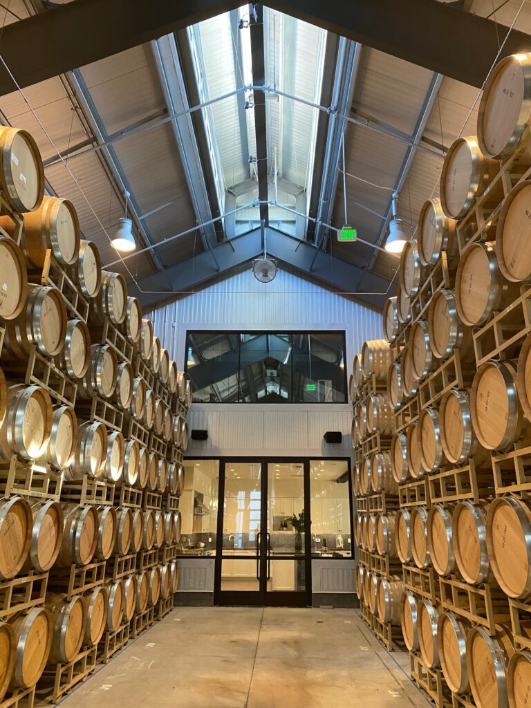 Long aisle inside a winery barrel room with stacked oak wine barrels on both sides and a skylit ceiling above.