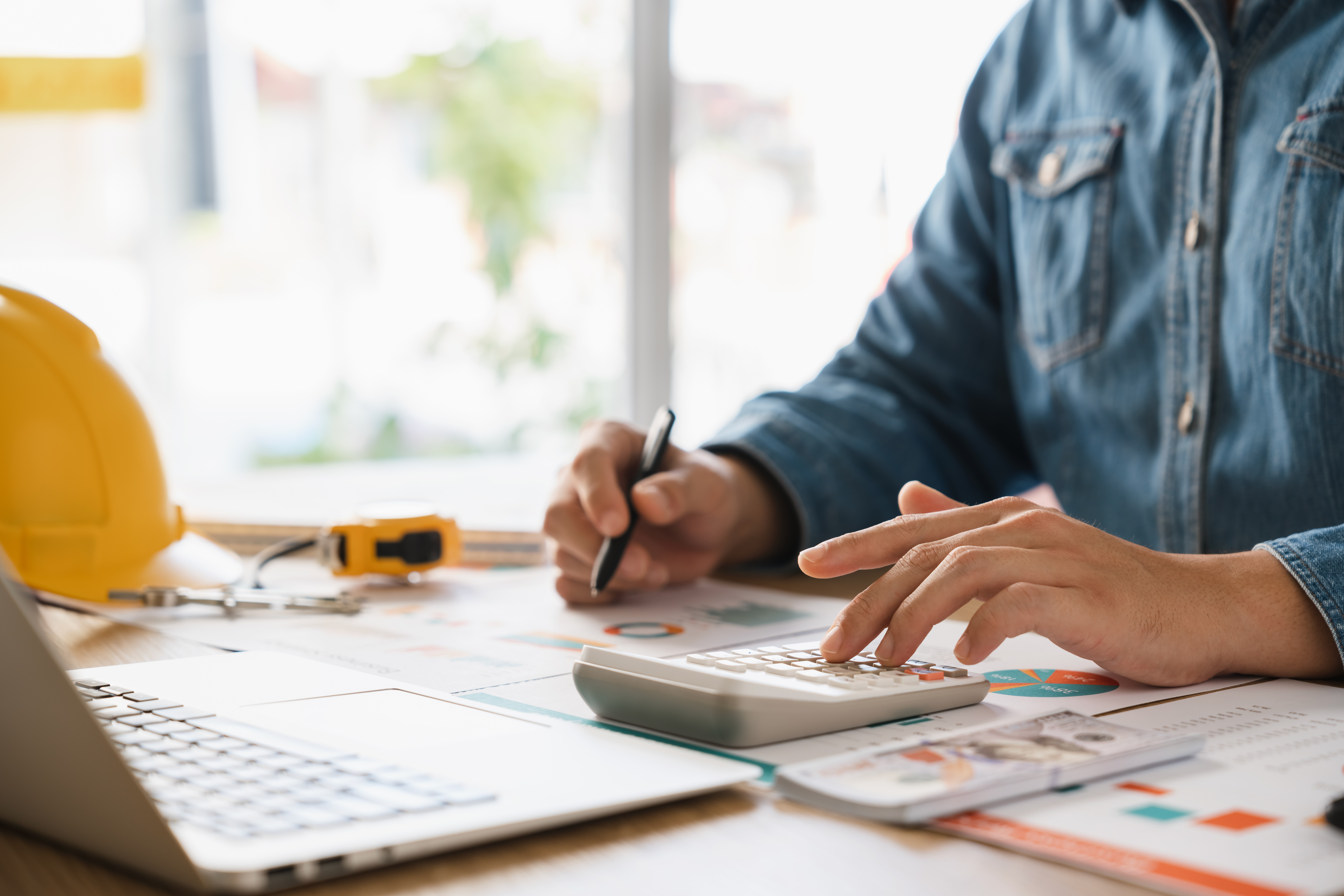 Close‑up of a plant manager in a denim shirt using a calculator and pen at a desk, with a laptop, project charts, yellow hard hat, and tape measure in view illustrating cost estimation work.
